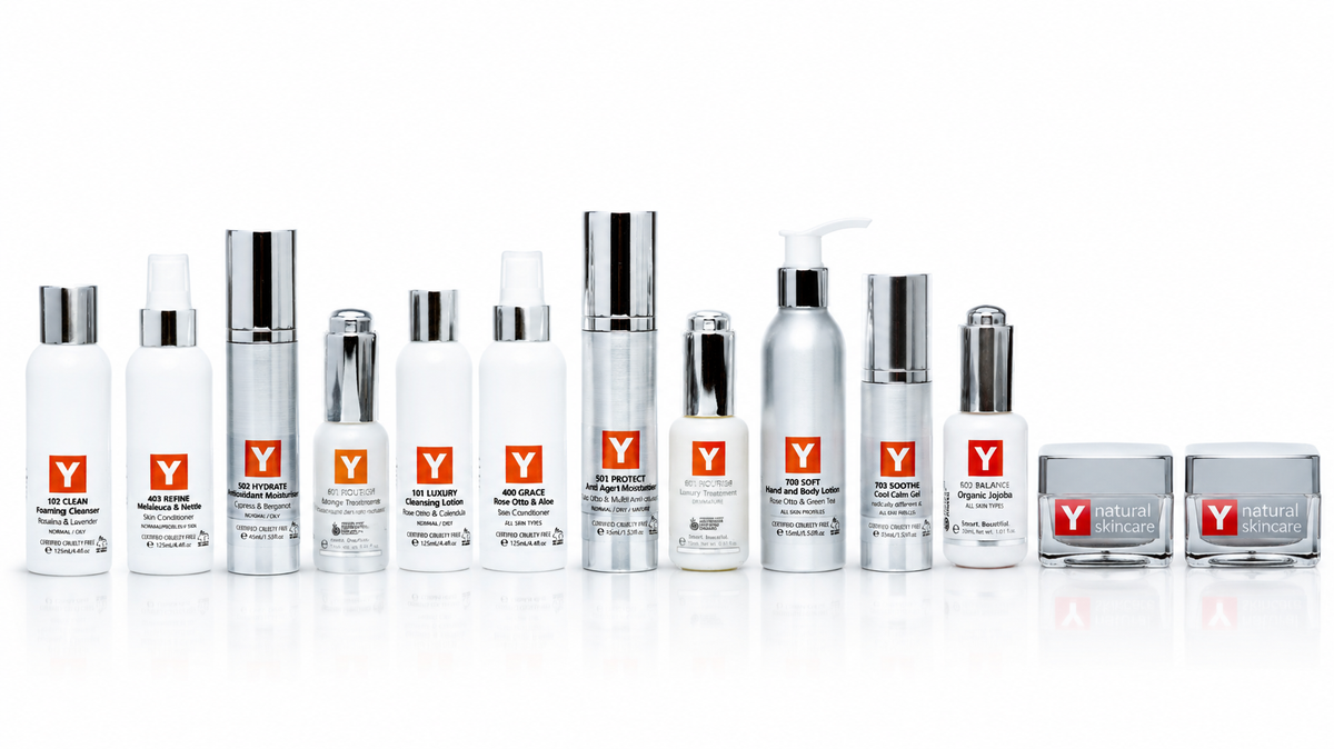A lineup of Y natural skincare products on a reflective white surface, featuring white and silver bottles, jars, and containers with a signature red Y logo.